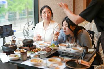 In a kalbi barbecue restaurant at a sunlit window table, two Korean women in their twenties in casual attire enjoy grilled meat and side dishes under soft spring light. Seoul, Jung District, spring.