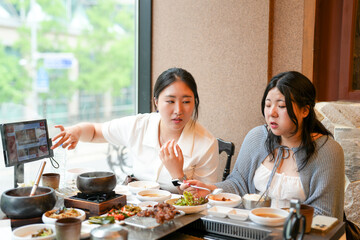 In a kalbi barbecue restaurant at a sunlit window table, two Korean women in their twenties in...
