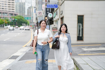 Two Korean women in their twenties in casual clothes walk along a boulevard sidewalk, hands on...