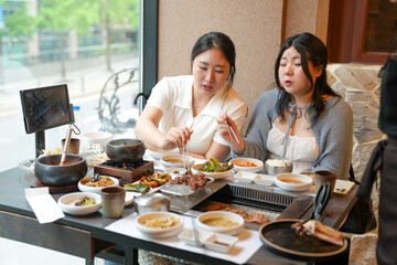 In a kalbi barbecue restaurant at a sunlit window table, two Korean women in their twenties in casual attire enjoy grilled meat and side dishes under soft spring light. Seoul, Jung District, spring.