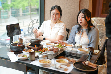 In a kalbi barbecue restaurant at a sunlit window table, two Korean women in their twenties in...
