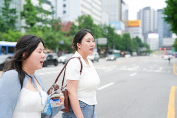 Two Korean women in their twenties in casual clothes walk across a crosswalk on a wide boulevard,...