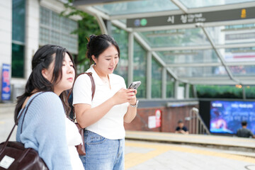 A Korean woman couple in their twenties in casual clothes stand by a stairway entrance to the...