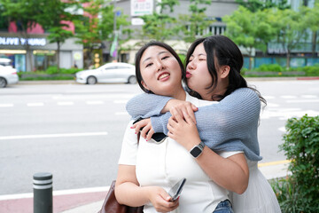 Two Korean women couple in their twenties in casual clothes embrace on a busy boulevard, leaning...