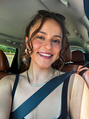 A beautiful well-dressed woman with her hair and make up done takes a selfie in a car during a sunny day.