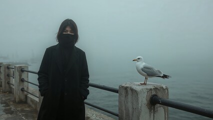 A person in dark clothing stands near a seagull on a foggy day by the water.