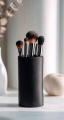 A black cylindrical makeup brush holder containing several brushes of varying sizes and shapes sits on a white surface, with a blurred background featuring a vase and dried plant