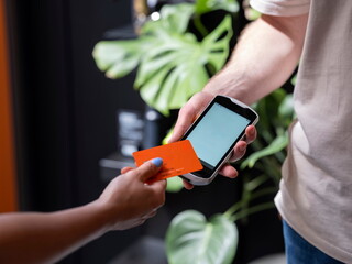 Person making contactless payment using orange card and smartphone in modern indoor setting with plants