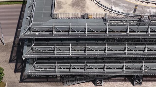 Elevated view of a modern multi-tier parking facility with metal ramps and a riverside promenade featuring cyclists and urban greenery.