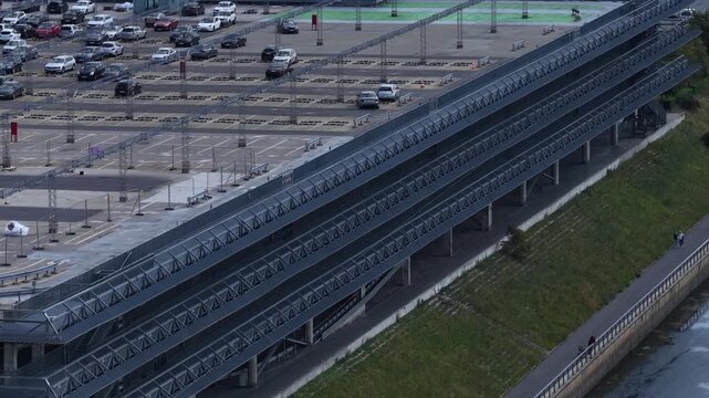 Elevated view of a modern multi-tier parking facility with metal ramps and a riverside promenade featuring cyclists and urban greenery.