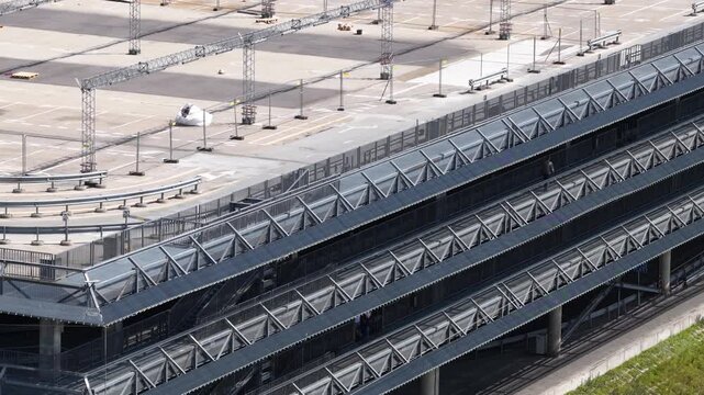 Elevated view of a modern multi-tier parking facility with metal ramps and a riverside promenade featuring cyclists and urban greenery.