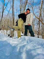 A couple takes a picture together snowshoeing in a snowy forest. The woman has her leg up with their dog in the background during the winter.