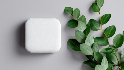 A white, square, textured cosmetic compact sits beside a sprig of eucalyptus leaves on a gray background