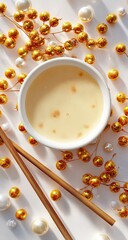 Creamy pale yellow liquid in a small white bowl, surrounded by golden and white beads and light wooden chopsticks on a white surface