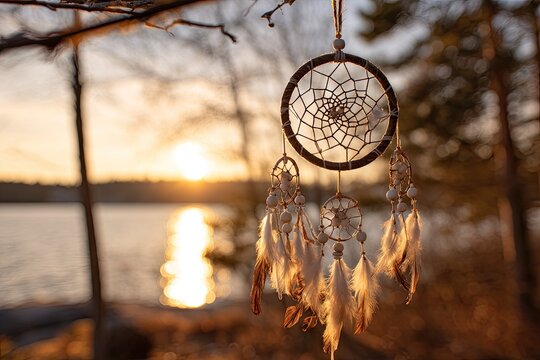 Sunset-lit dreamcatcher hangs from a tree branch overlooking a calm lake, its soft feathers catching the golden light