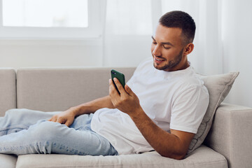 Obraz premium Young man relaxing on a couch, smiling while using a smartphone in a bright, cozy living room, showcasing modern lifestyle and technology trends.