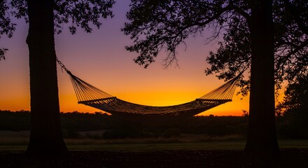 Obraz premium Hammock silhouette between trees against a vibrant sunset sky with orange and purple hues in the background