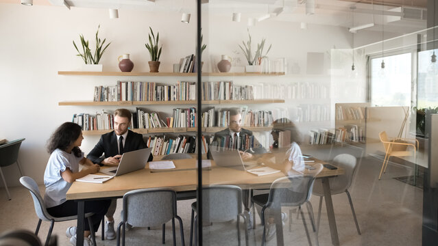 In modern conference room two multiethnic skilled colleagues solving joint business-related matters, sitting at desk with laptop and papers, leading formal conversation, share solution, and strategy