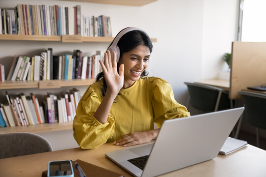 Positive Indian female teacher or counselor wear headphones sits at desk with laptop greeting student or client start online videocall class lecture using modern wireless technology. Virtual meeting - Powered by Adobe