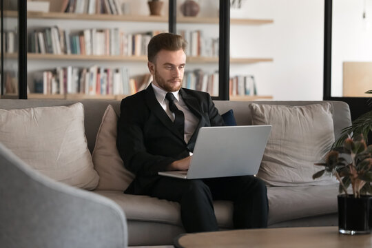 Businessman dressed in suit sit on sofa in coworking, focused on working with laptop, preparing business strategies, reports, or proposals, responds to e-mails, reviewing documents, or financial data