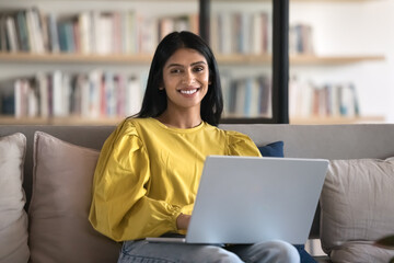 Portrait of pretty Indian woman sit on comfortable sofa with laptop, smile, look at camera, feel satisfied with workflow in modern coworking office, preparing tasks, working remotely. Freelance, study