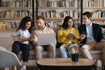 Four students or employees resting in coworking space lobby or library with diverse devices, discussing new business application or educations platforms, share news enjoy break. Modern wireless tech