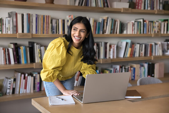 Happy Indian woman distracted from working on laptop, looks aside with smile, enjoy work results, achievements and productive workday in modern creative workspace. Workflow, aspirations, motivation