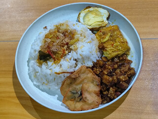 A dish of mixed rice with side dishes of orek tempeh, salted egg, perkedel, and yellow spiced egg, served on a white melamine plate on a wooden table. Traditional Indonesian food mixed rice.
