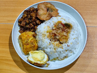A dish of mixed rice with side dishes of orek tempeh, salted egg, perkedel, and yellow spiced egg, served on a white melamine plate on a wooden table. Traditional Indonesian food mixed rice.