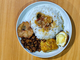 A dish of mixed rice with side dishes of orek tempeh, salted egg, perkedel, and yellow spiced egg, served on a white melamine plate on a wooden table. Traditional Indonesian food mixed rice.