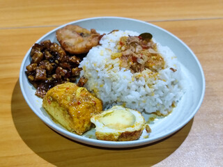 A dish of mixed rice with side dishes of orek tempeh, salted egg, perkedel, and yellow spiced egg, served on a white melamine plate on a wooden table. Traditional Indonesian food mixed rice.
