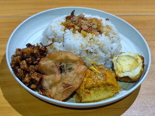 A dish of mixed rice with side dishes of orek tempeh, salted egg, perkedel, and yellow spiced egg, served on a white melamine plate on a wooden table. Traditional Indonesian food mixed rice.
