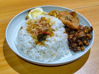 A dish of mixed rice with side dishes of orek tempeh, salted egg, perkedel, and yellow spiced egg, served on a white melamine plate on a wooden table. Traditional Indonesian food mixed rice.