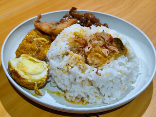 A dish of mixed rice with side dishes of orek tempeh, salted egg, perkedel, and yellow spiced egg, served on a white melamine plate on a wooden table. Traditional Indonesian food mixed rice.