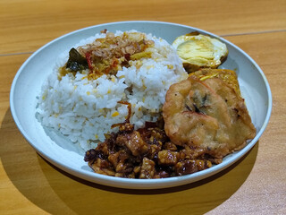 A dish of mixed rice with side dishes of orek tempeh, salted egg, perkedel, and yellow spiced egg, served on a white melamine plate on a wooden table. Traditional Indonesian food mixed rice.