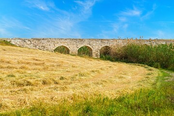 An old stone aqueduct bridge looms over a field of dry grass under a clear blue sky. This serene landscape captures a peaceful moment, perfect for travel or historical themes.