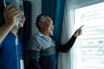 senior man patient standing near window looks outside with his elderly nurse assistant. home...