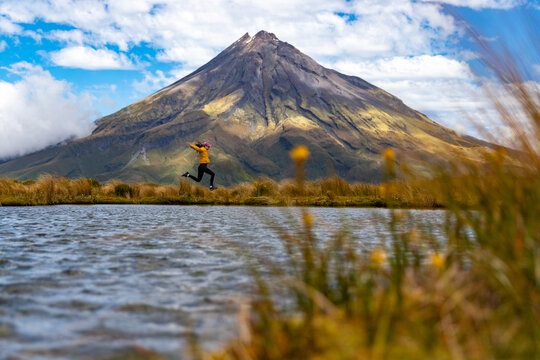 Woman jumps high by Pouakai Tarn with the mighty Mount Taranaki in background, Egmont National Park, North Island, New Zealand