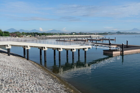 A clean and modern pier stretches over calm waters at Sanur Port, Bali, with mountains and coastal views in the background.