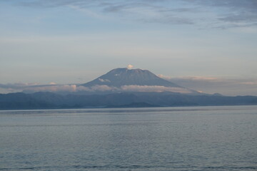 Fototapeta premium A majestic view of Mount Agung rising above clouds, seen from across the calm ocean waters of Nusa Penida, Bali at sunrise.