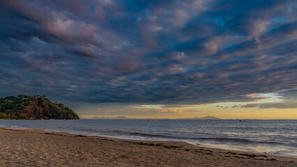 Sunset on a tropical island. Ocean waves are foaming on a sandy beach. Blue clouds are highlighted in pink. The sky is orange on the horizon. A hill in the distance. Madagascar. Nosy Be  