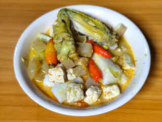 Traditional Indonesian vegetable lontong dish. Lontong opor ayam with sliced chayote, carrots, tofu and sliced red chilies, served in a white ceramic bowl on a wooden table.