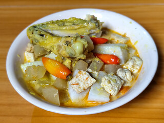Traditional Indonesian vegetable lontong dish. Lontong opor ayam with sliced chayote, carrots, tofu and sliced red chilies, served in a white ceramic bowl on a wooden table.
