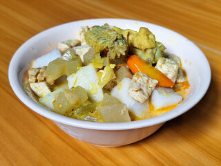 Traditional Indonesian vegetable lontong dish. Lontong opor ayam with sliced chayote, carrots, tofu and sliced red chilies, served in a white ceramic bowl on a wooden table.