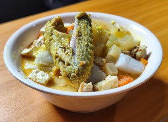 Traditional Indonesian vegetable lontong dish. Lontong opor ayam with sliced chayote, carrots, tofu and sliced red chilies, served in a white ceramic bowl on a wooden table.