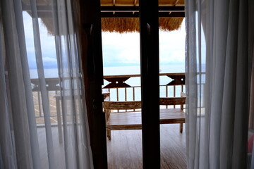 A tranquil view through sheer white curtains revealing a bamboo balcony with a bench, overlooking...