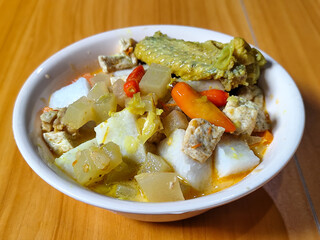 Traditional Indonesian vegetable lontong dish. Lontong opor ayam with sliced chayote, carrots, tofu and sliced red chilies, served in a white ceramic bowl on a wooden table.