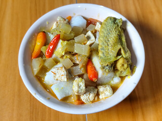 Traditional Indonesian vegetable lontong dish. Lontong opor ayam with sliced chayote, carrots, tofu and sliced red chilies, served in a white ceramic bowl on a wooden table.