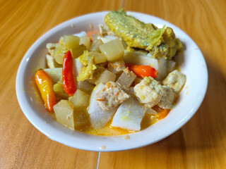 Traditional Indonesian vegetable lontong dish. Lontong opor ayam with sliced chayote, carrots, tofu and sliced red chilies, served in a white ceramic bowl on a wooden table.