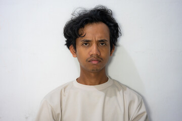 A young Indonesian-asian man with a serious expression and messy hair, standing against a white wall. Perfect for portraying emotions, portraits, or lifestyle projects.